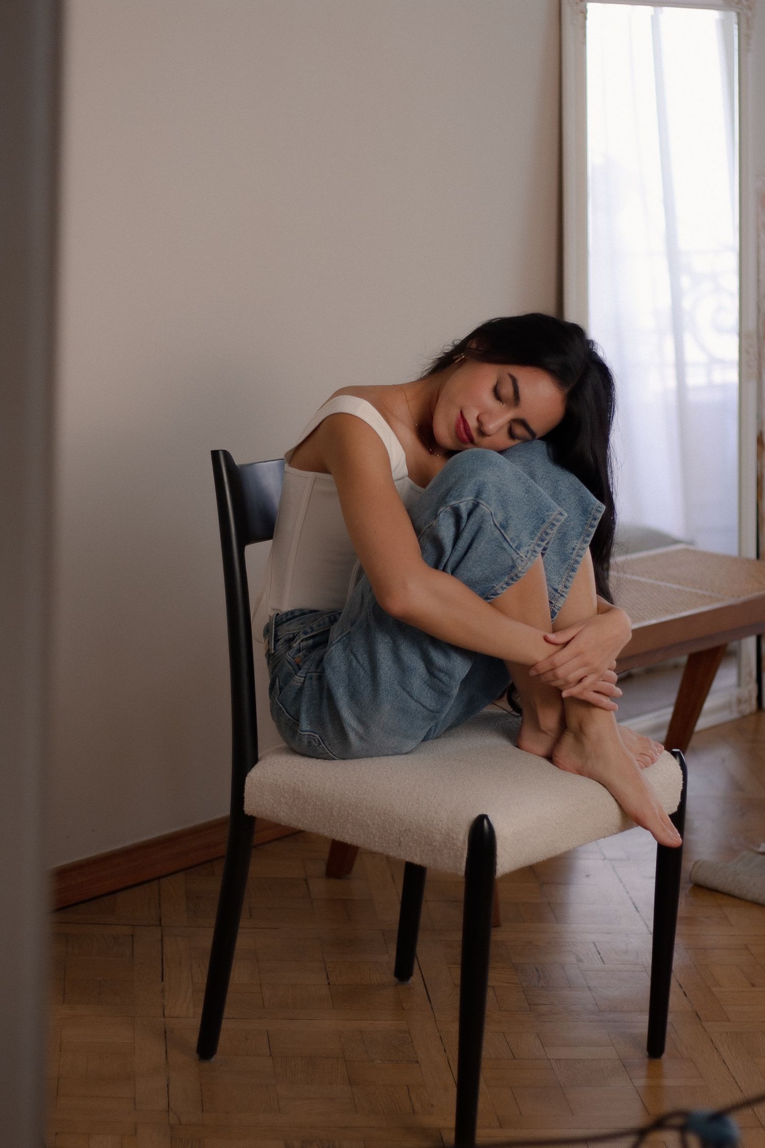 Young brunette woman sitting curled up on a chair hugging her knees in a cozy indoor setting.