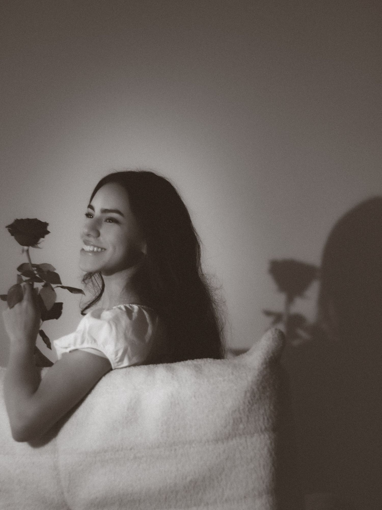 Smiling young woman holding a single rose with its shadow cast on the wall behind her.