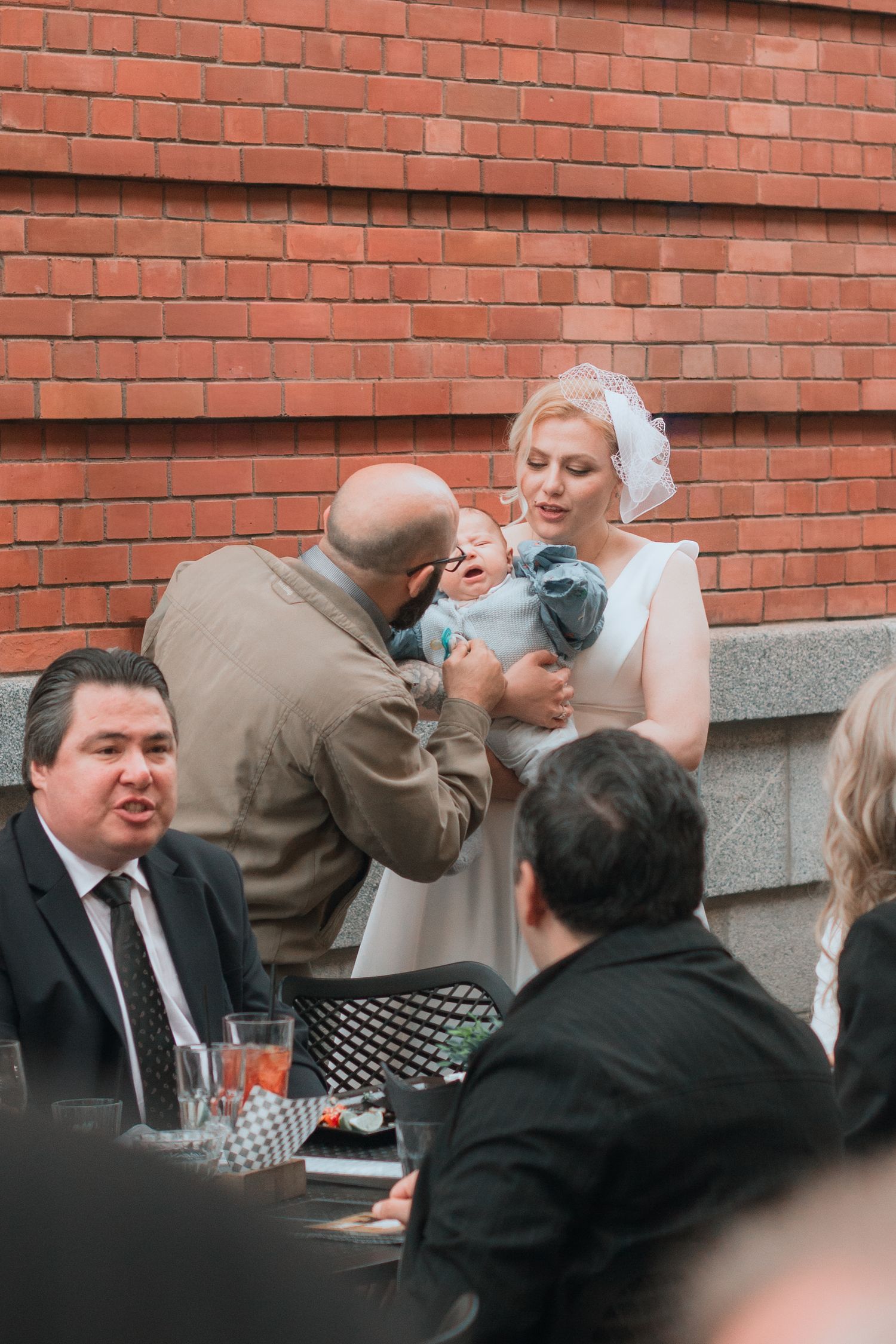 Bride in white dress holds baby while bald groom interacts with infant at outdoor wedding reception.
