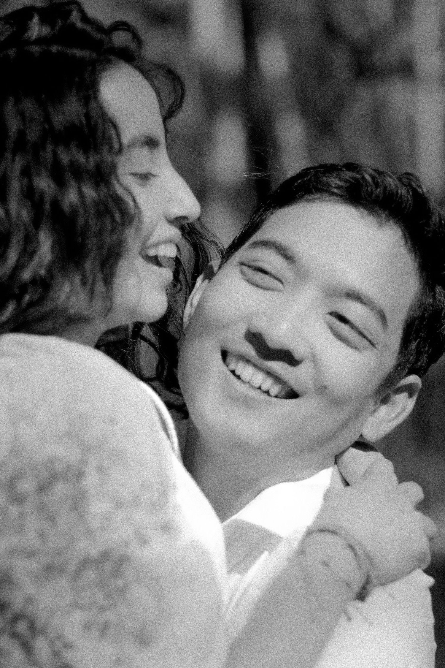 Black and white photo of laughing couple embracing closely outdoors in a bright natural setting.