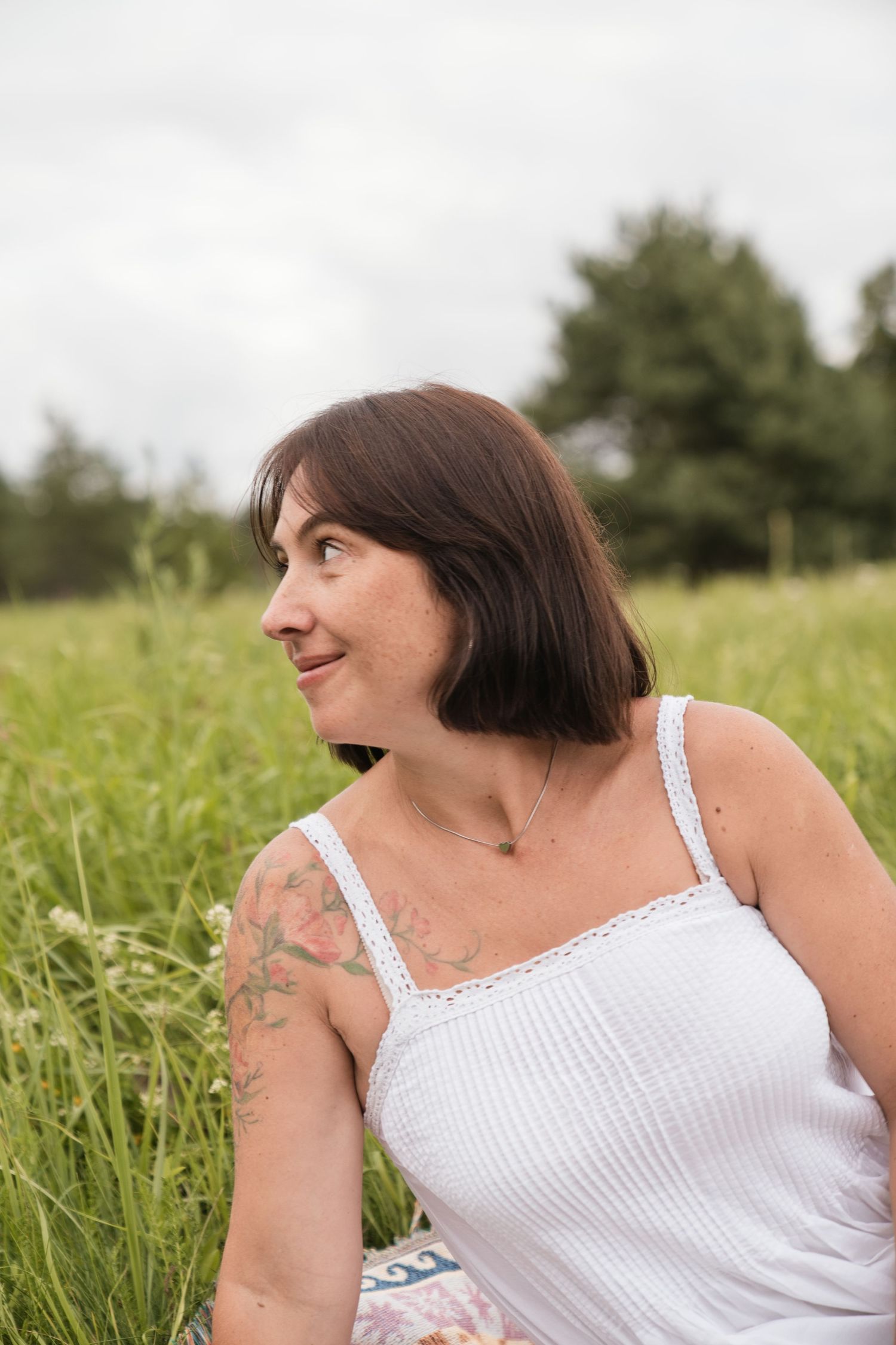 A smiling brunette woman with a tattoo sits in a lush green meadow wearing a white sundress on a cloudy day.