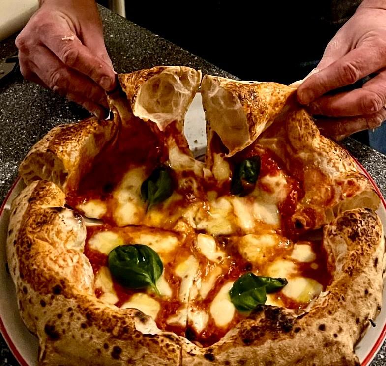 Hands pulling a slice of authentic Neapolitan margherita pizza with stretchy mozzarella, basil, and charred crust.