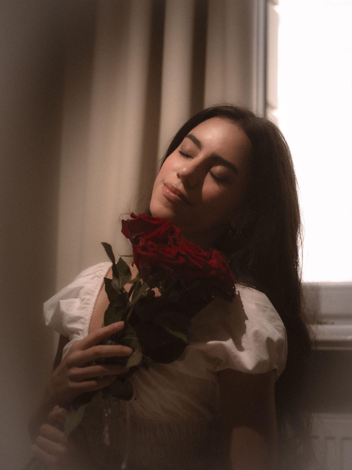 Young woman with long dark hair holding red roses with eyes closed near a curtained window.