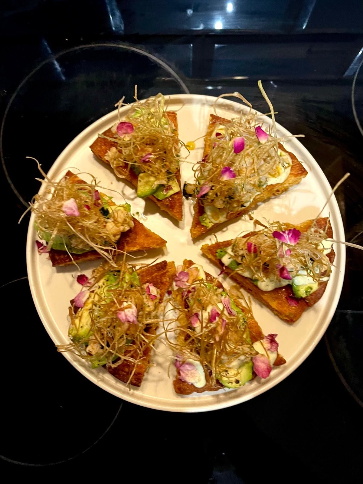 A round white plate with triangular focaccia slices garnished with avocado, rose petals, and crispy strands.