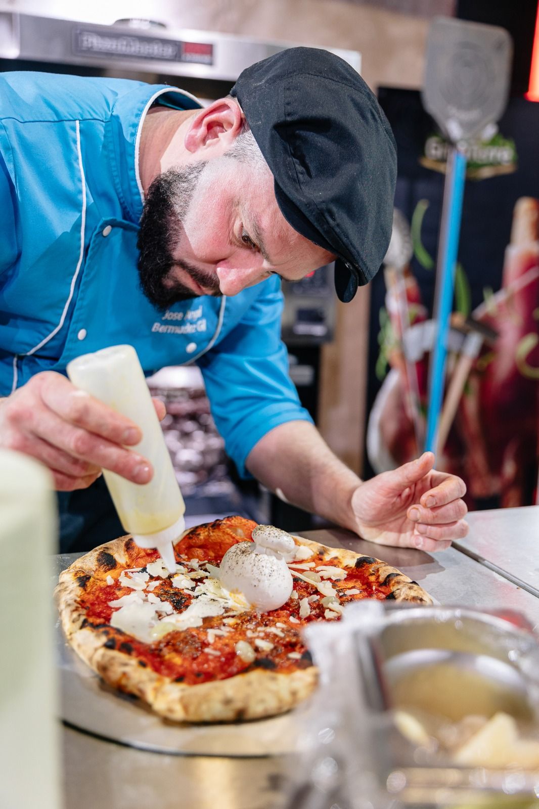 Focused male chef in blue uniform carefully garnishing a Neapolitan pizza with burrata at competition.