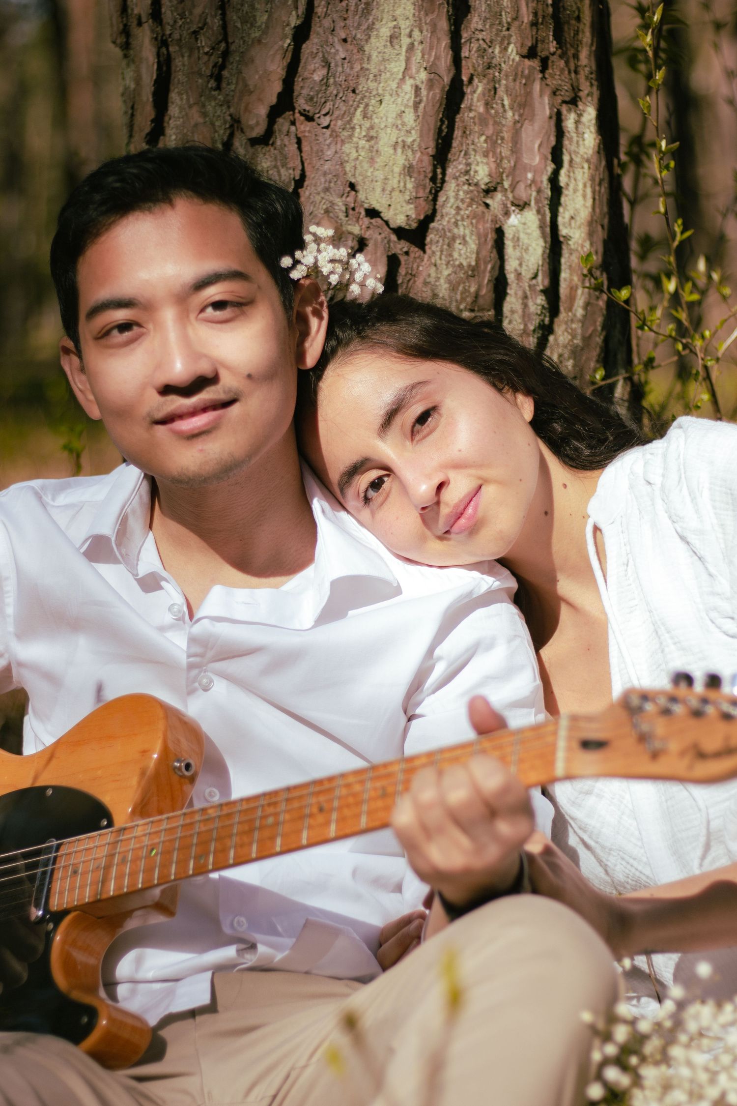 Couple sitting against a tree, man playing guitar while woman rests her head on his shoulder outdoors.