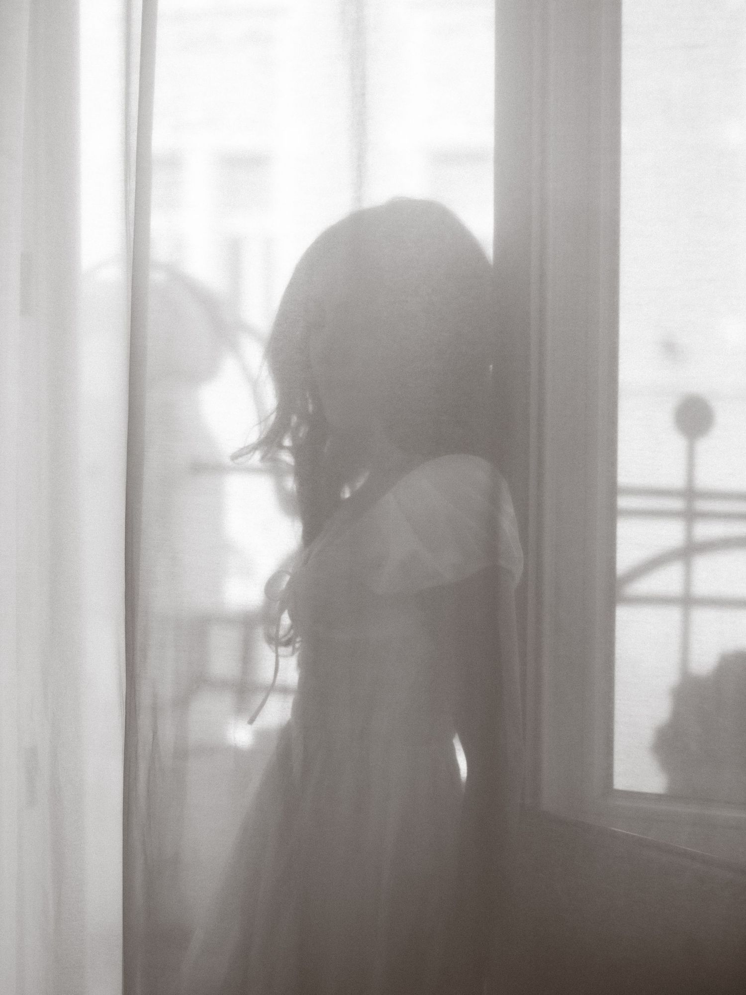 Silhouette of a woman in a dress standing by a bright window with sheer curtains in a dreamy black and white photo.