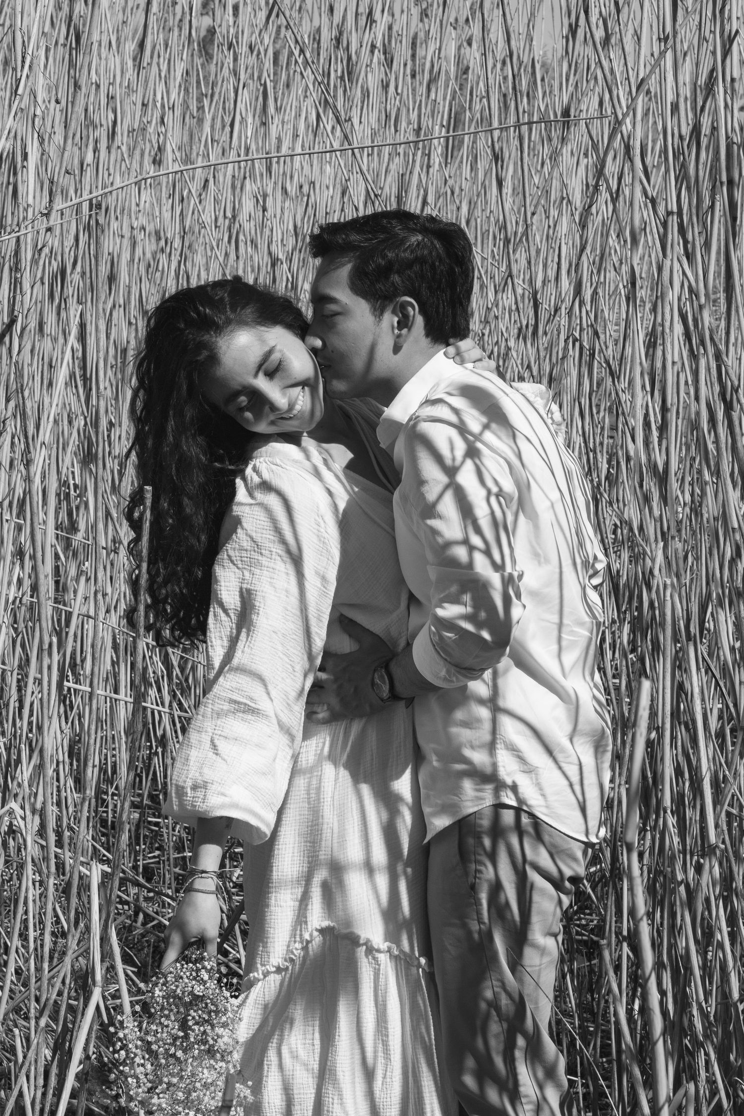 Man kissing woman on cheek in black and white photo among tall dry reeds.