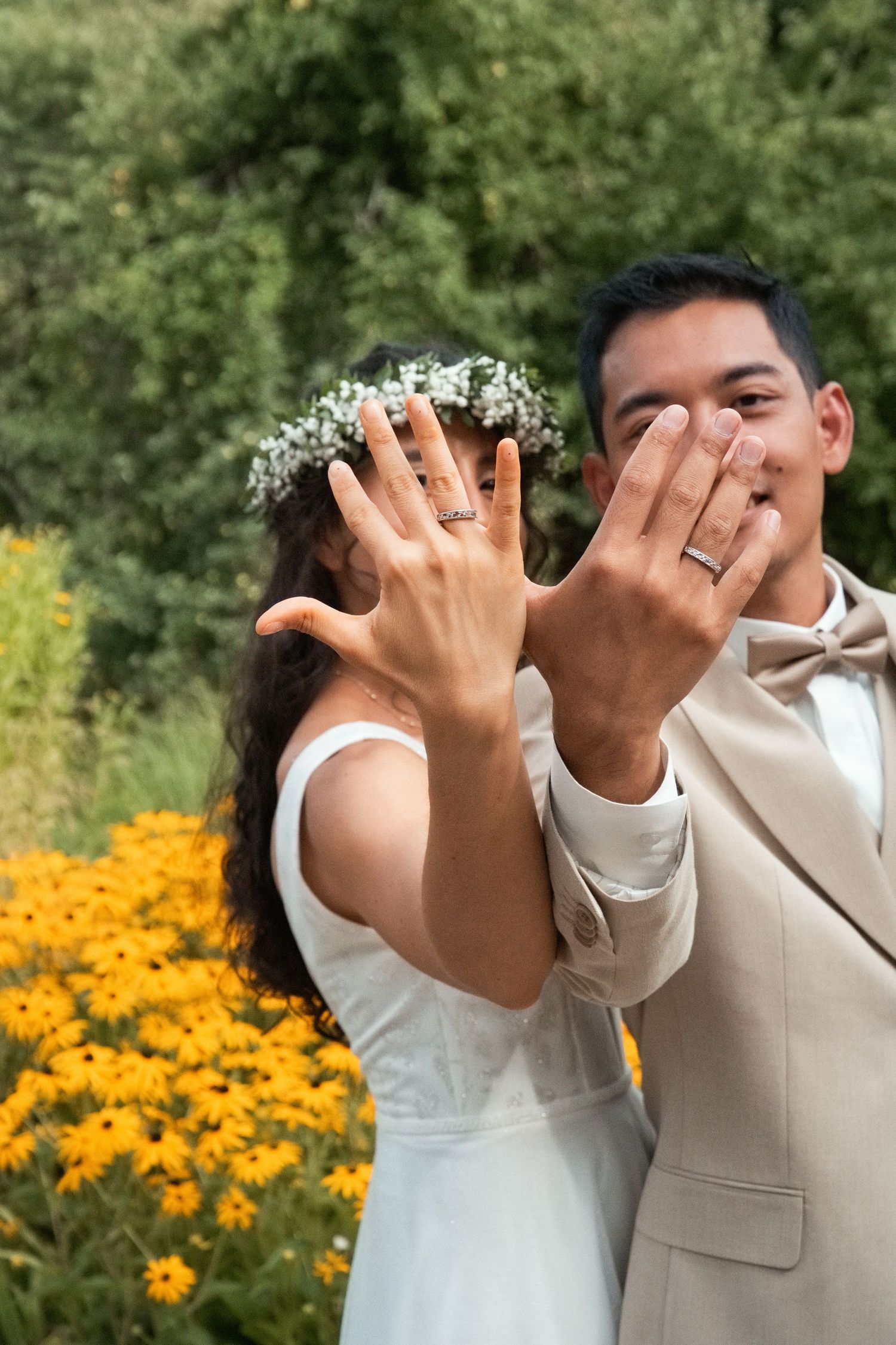 Newlyweds showing off wedding rings to camera with yellow flowers in background.
