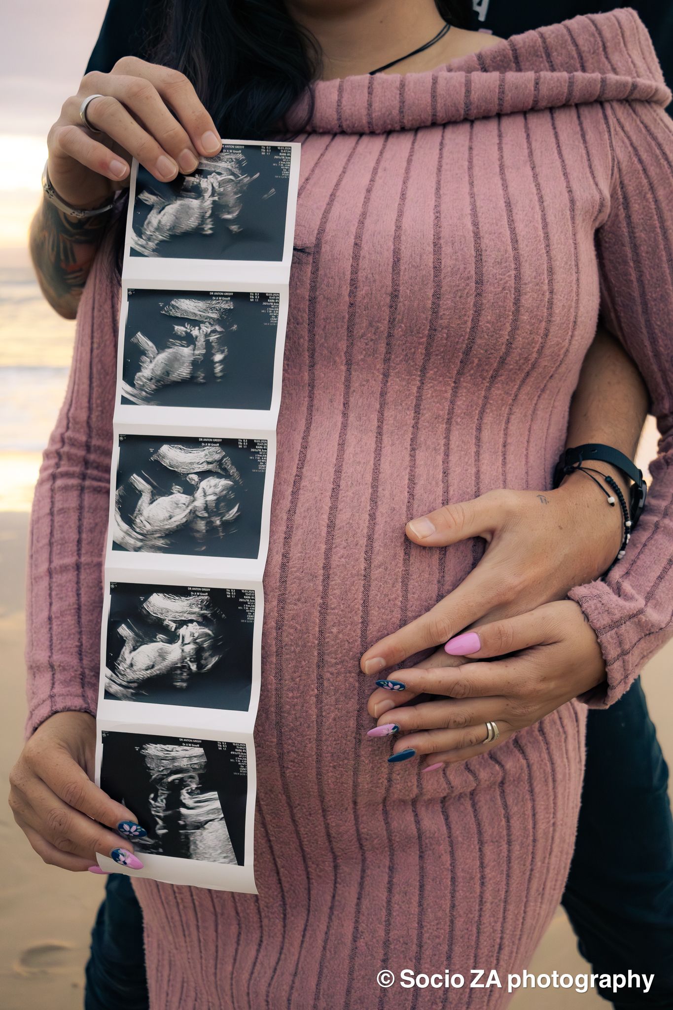 Sonogram in front of a woman standing on the beach while pregnant with her husband holding her stomach. Portrait in Jeffrey’s Bay.