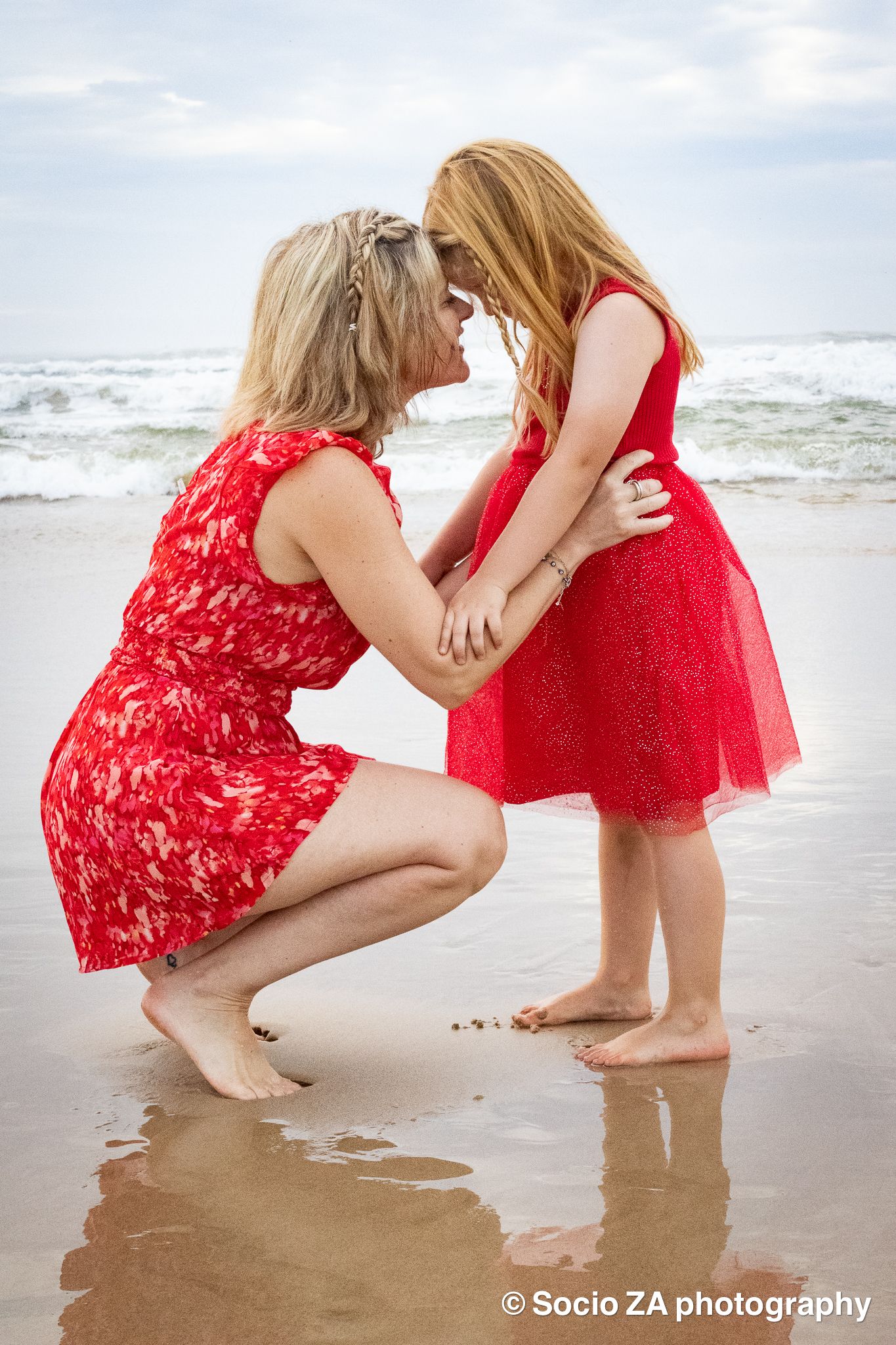 Mother and daughter touching faces in a sincere. moment of love while wearing red dresses on the beach in Jeffreys Bay Eastern Cape.