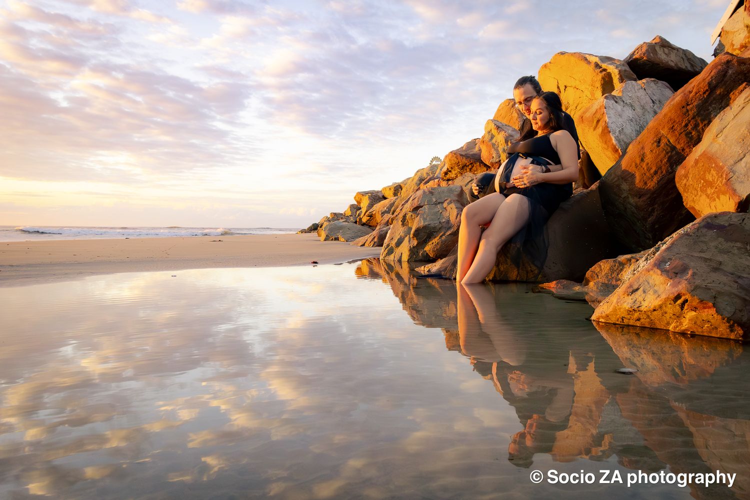 Maternity photo of a woman sitting on a rock at the beach while holding her stomach with her husband next to her. Sunrise Jeffreys Bay.