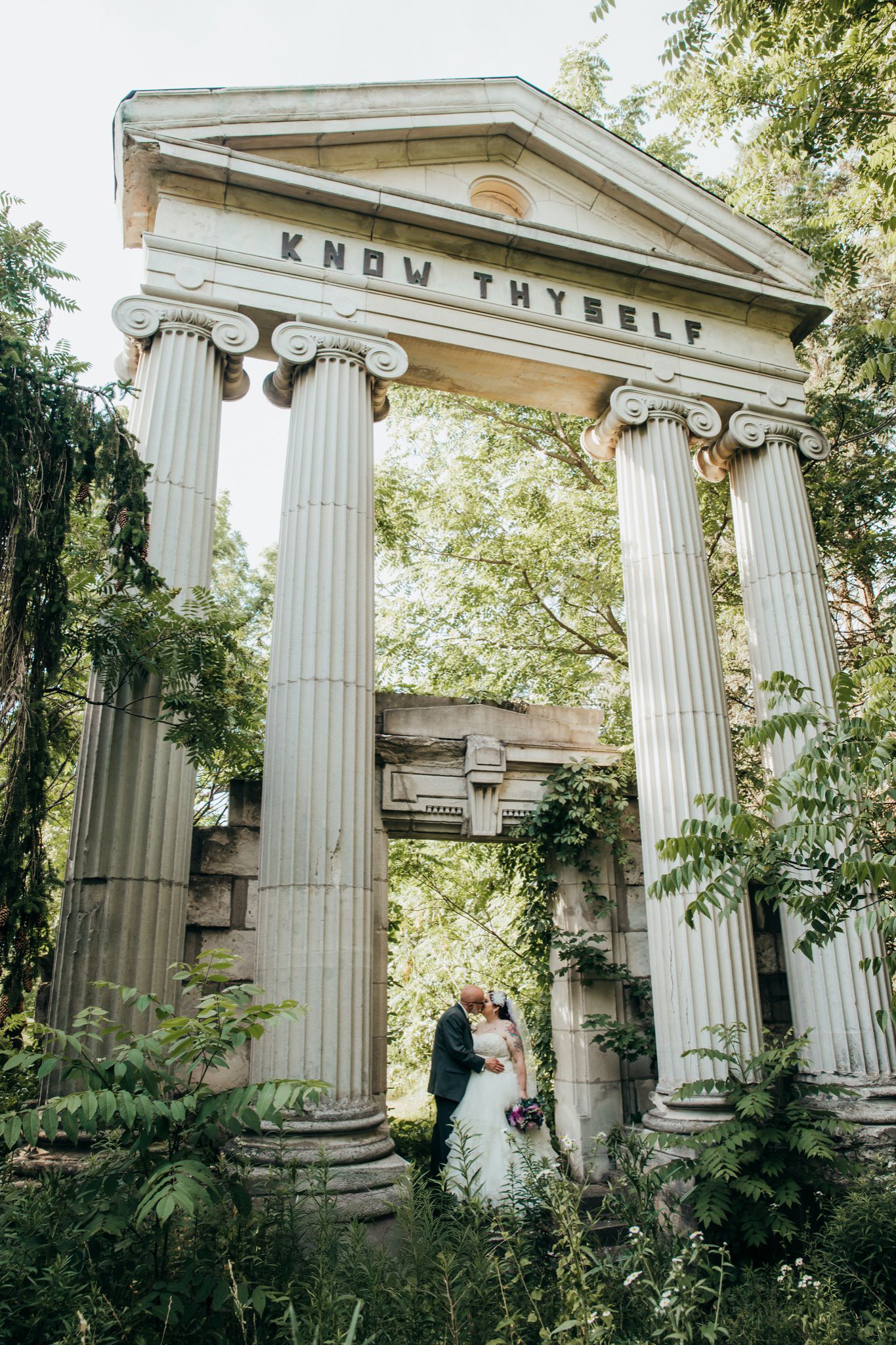 Wedding couple photographed among classical Greek columns in a garden setting.