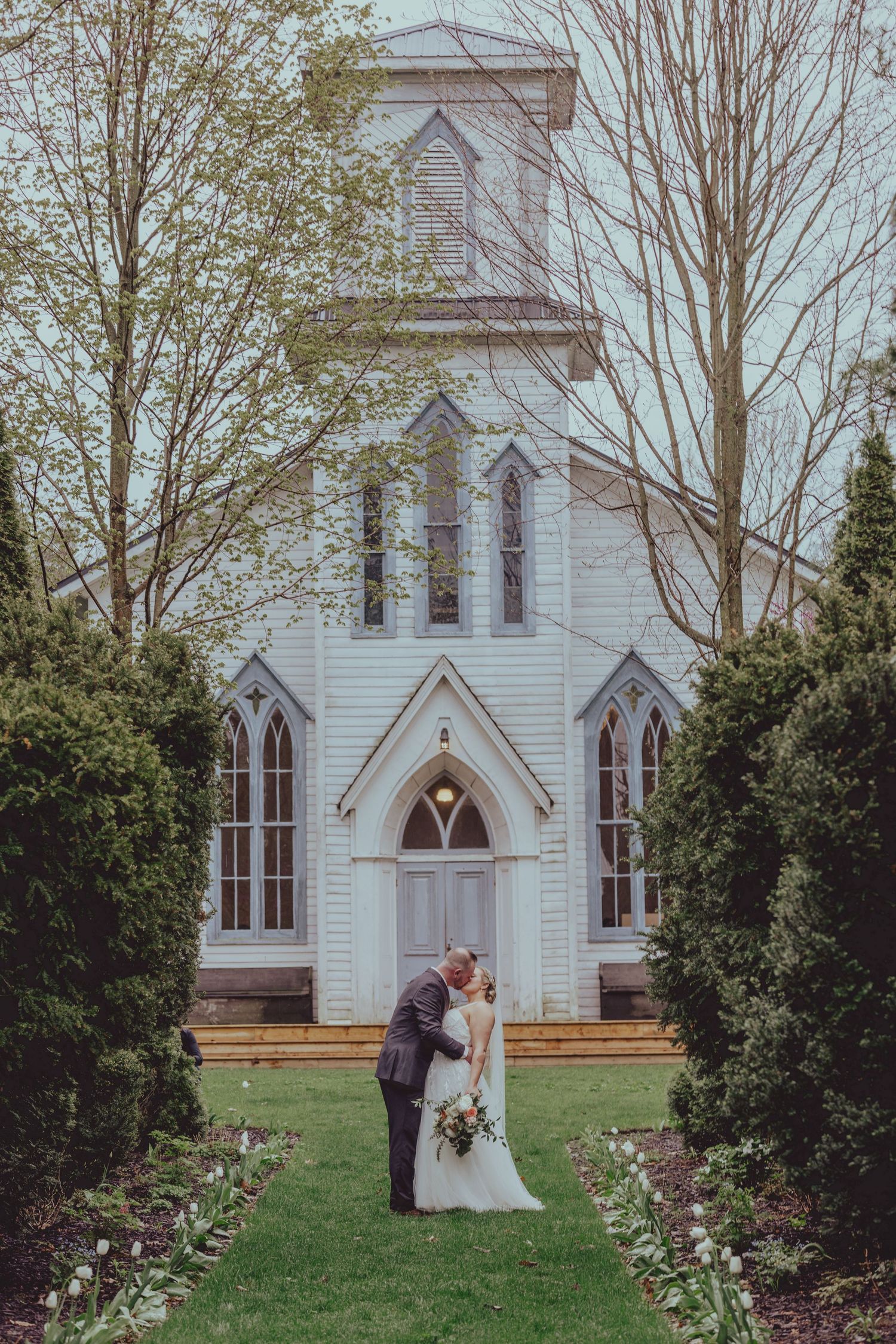 Wedding couple poses in front of white gothic revival church on grassy lawn.