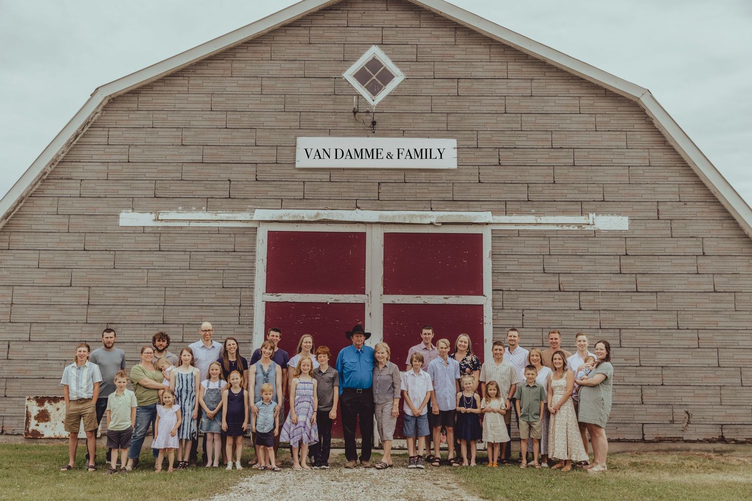 Large group poses in front of a tan brick barn building with red doors.