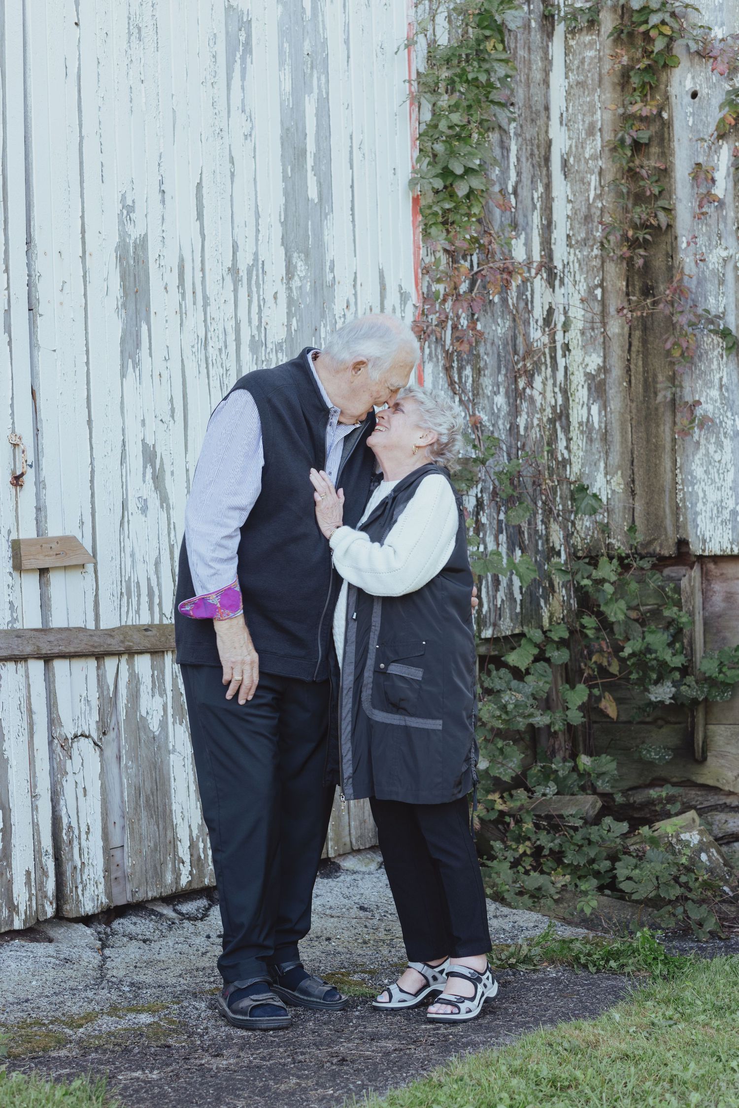Sweet candid moments captured against a weathered white barn wall with climbing vines.