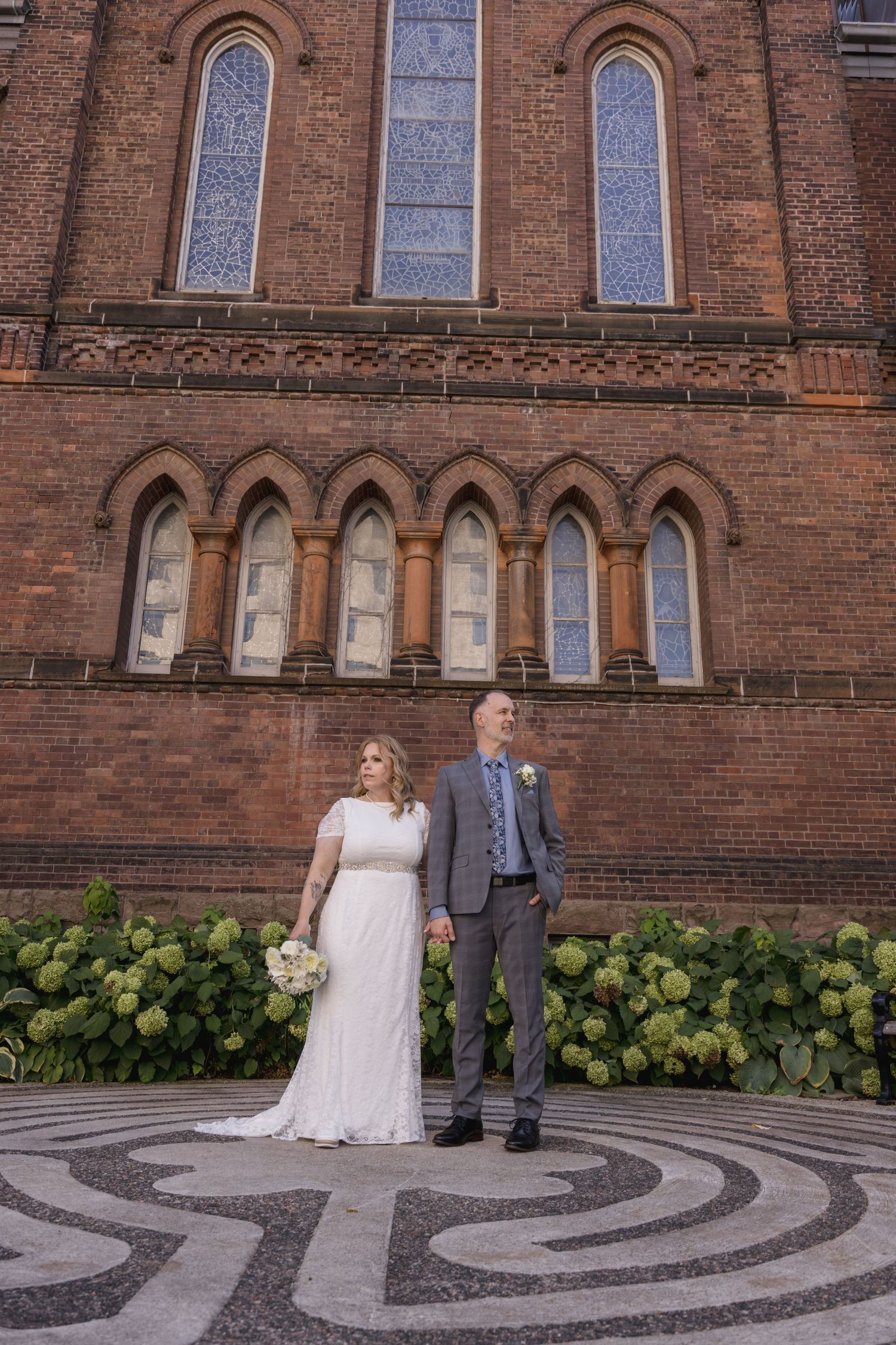 Wedding couple poses outside historic brick church with ornate architecture and hydrangea bushes.