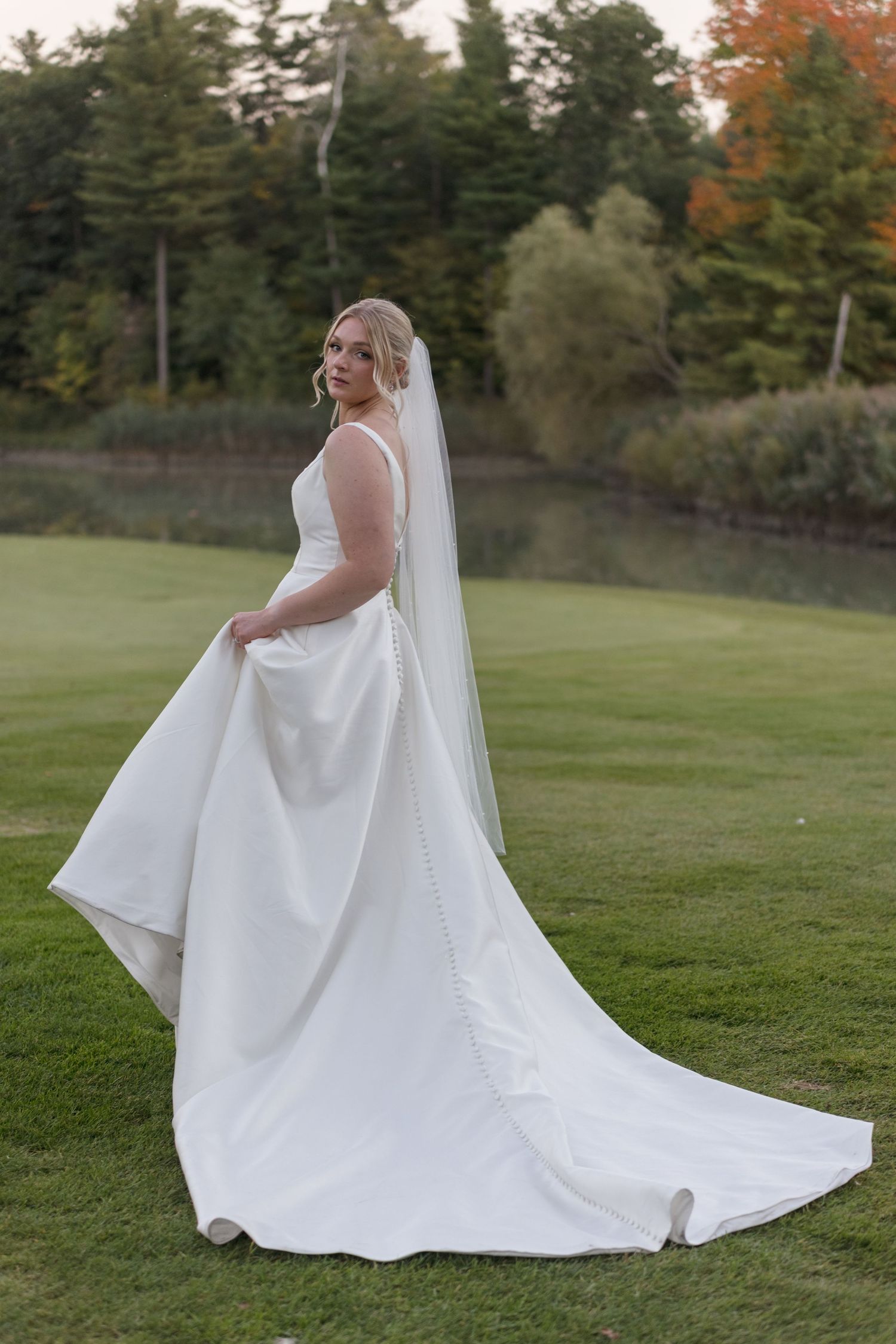 Bride in white satin wedding gown posing on grassy lawn with fall foliage in background.