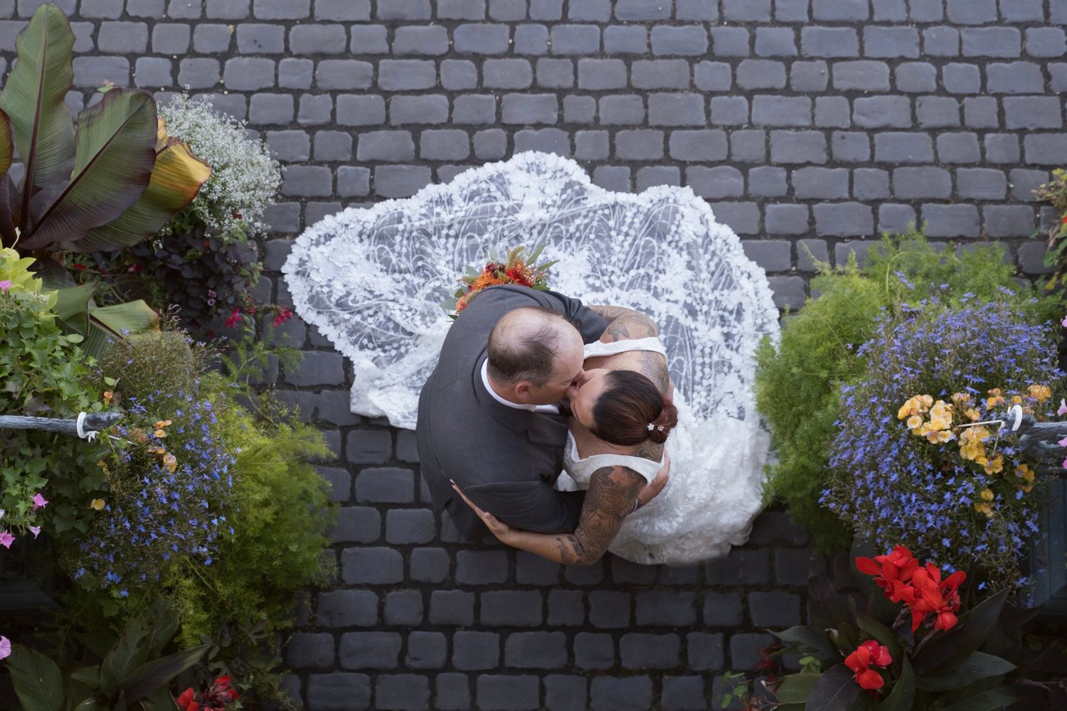 Aerial view of someone in a white lace wedding dress lying on cobblestone pavement surrounded by flowers.