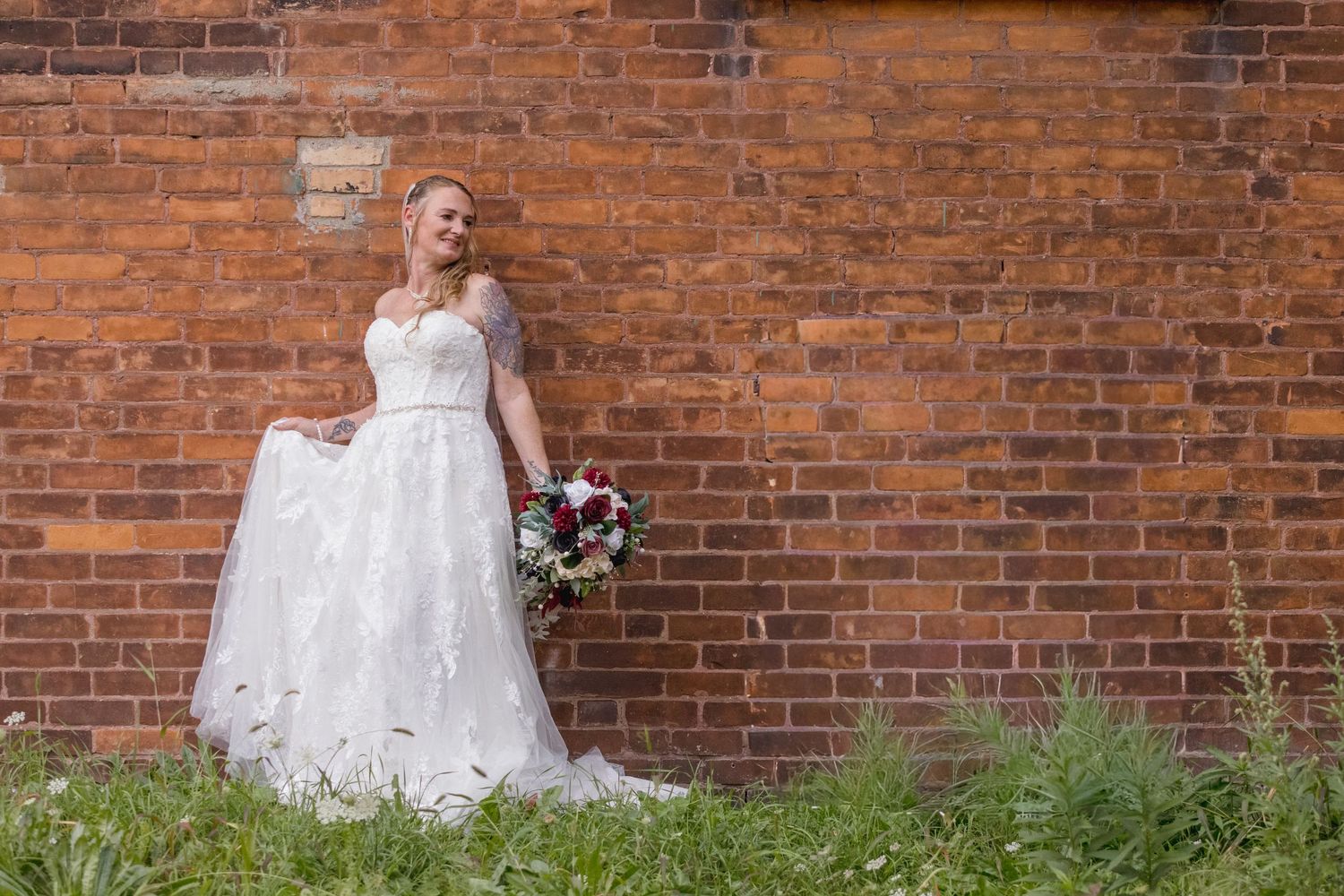 Bride in white wedding dress poses against rustic brick wall with greenery.