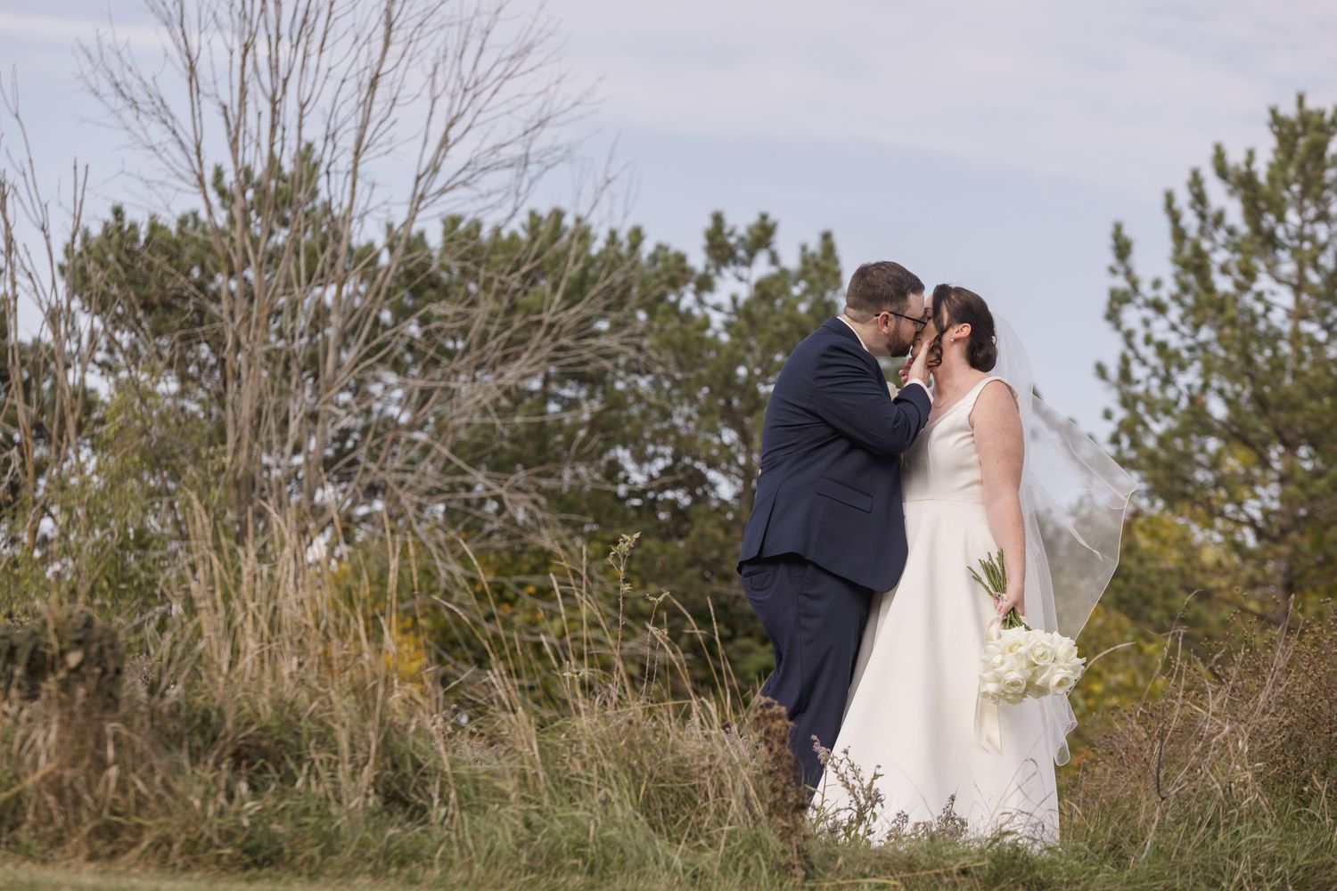 Newlyweds share an intimate moment against a natural landscape with trees.