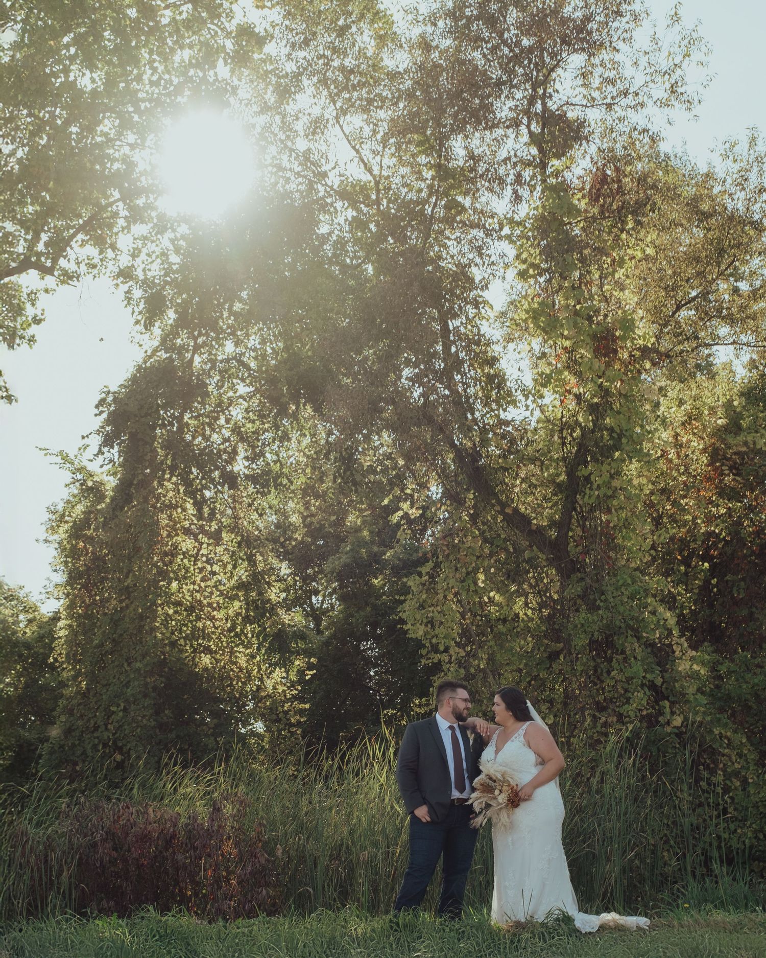 Wedding couple walking through a sunlit meadow with tall grass and trees.