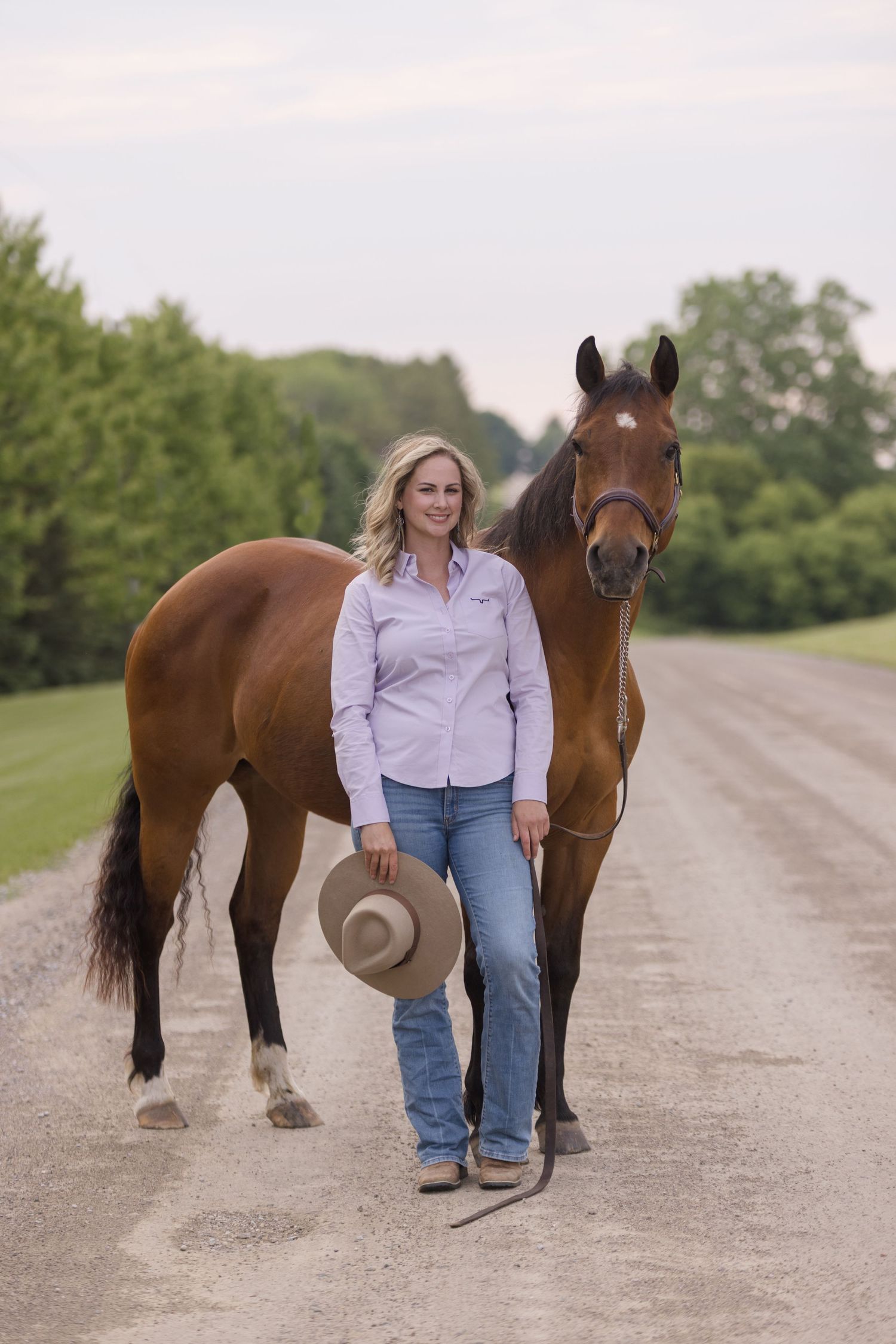 Person in purple shirt and jeans standing with brown horse on a country path.