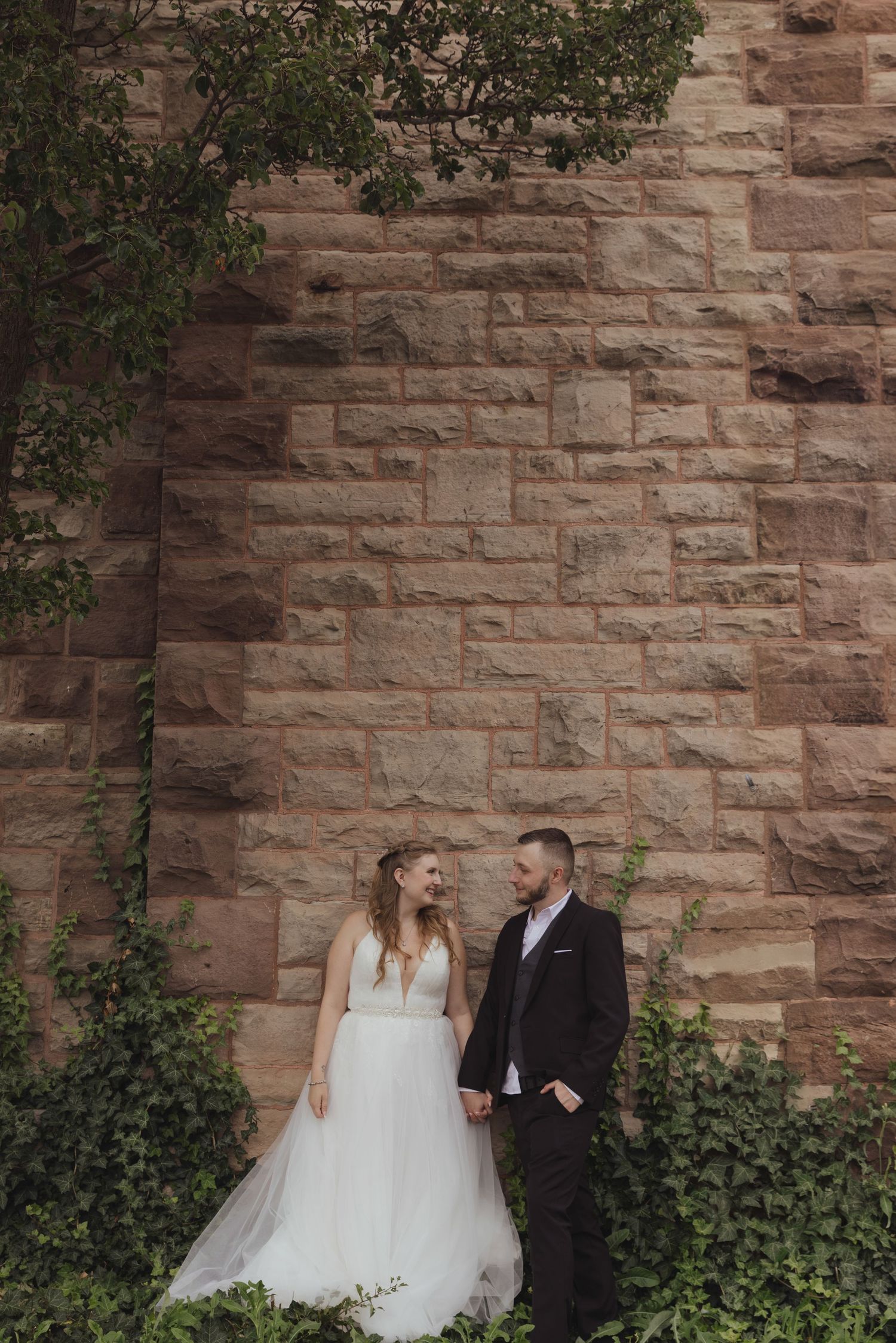 Couple in wedding attire posing against a rustic stone wall with ivy.