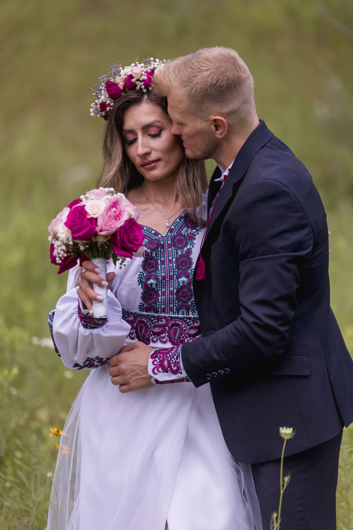 Wedding couple in traditional embroidered attire with pink floral crown and bouquet.