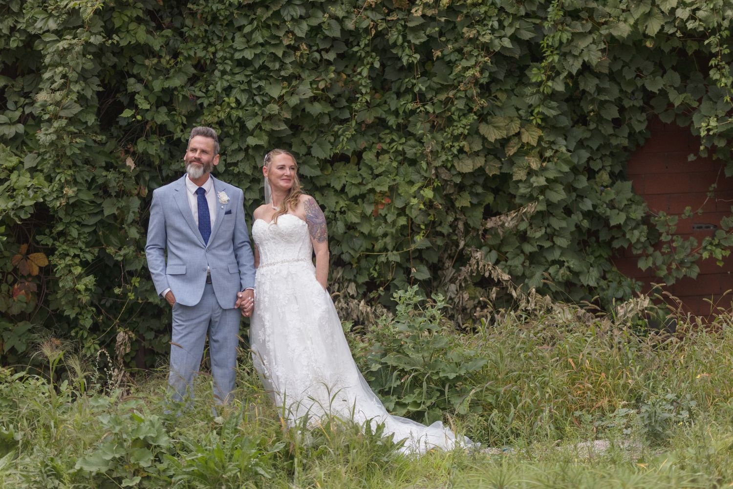 Couple sits together on ground against backdrop of lush green bushes and brick wall.