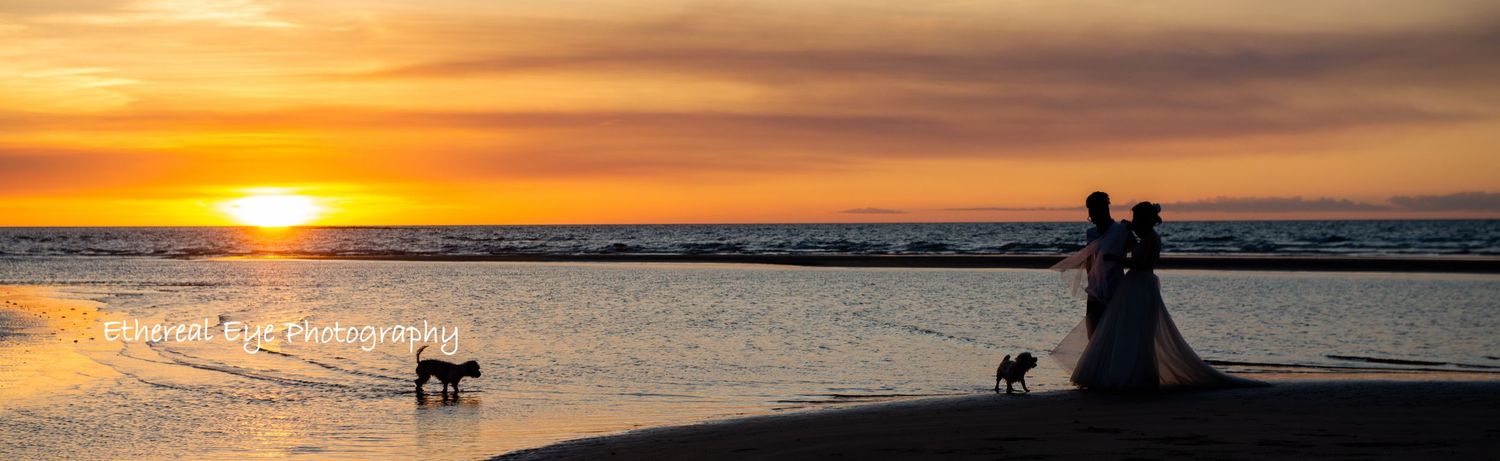 Panoramic view of a dramatic orange and purple sunset over the ocean with silhouettes in the foreground.
