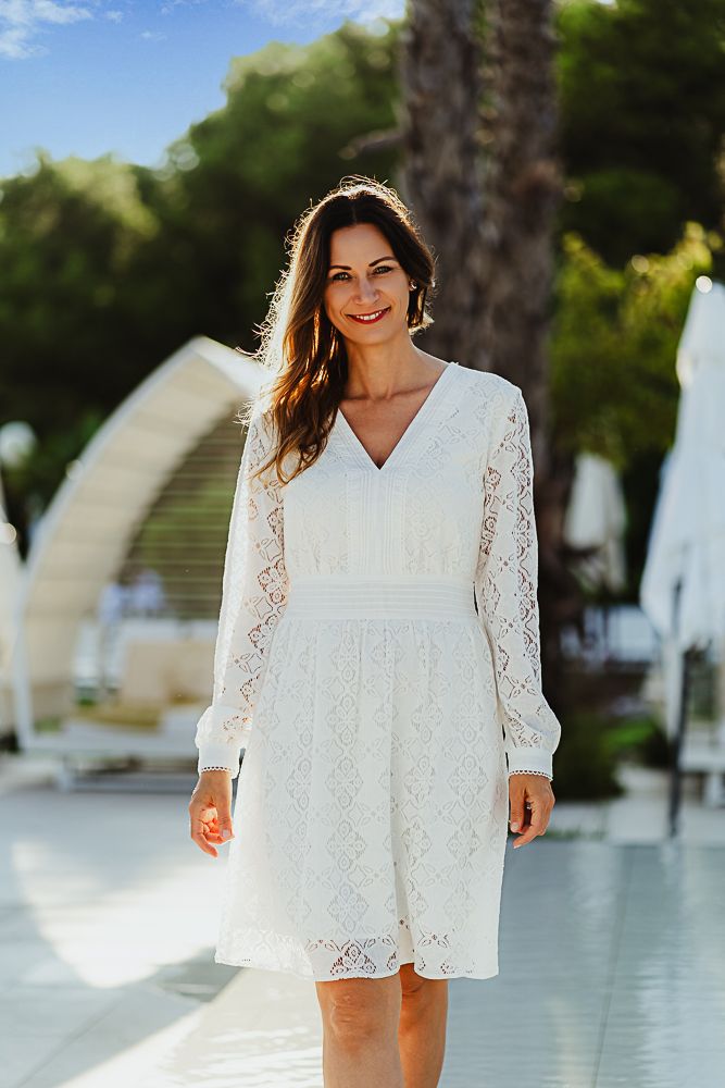 Woman in white lace dress standing by pool with palm trees in sunny outdoor setting.