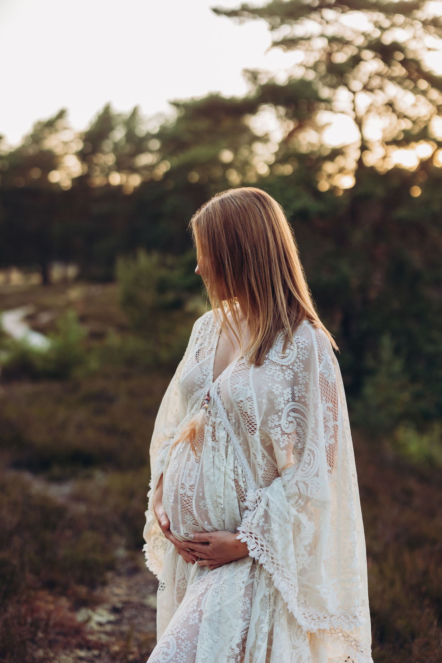 Woman in white lace dress standing among trees during golden hour.