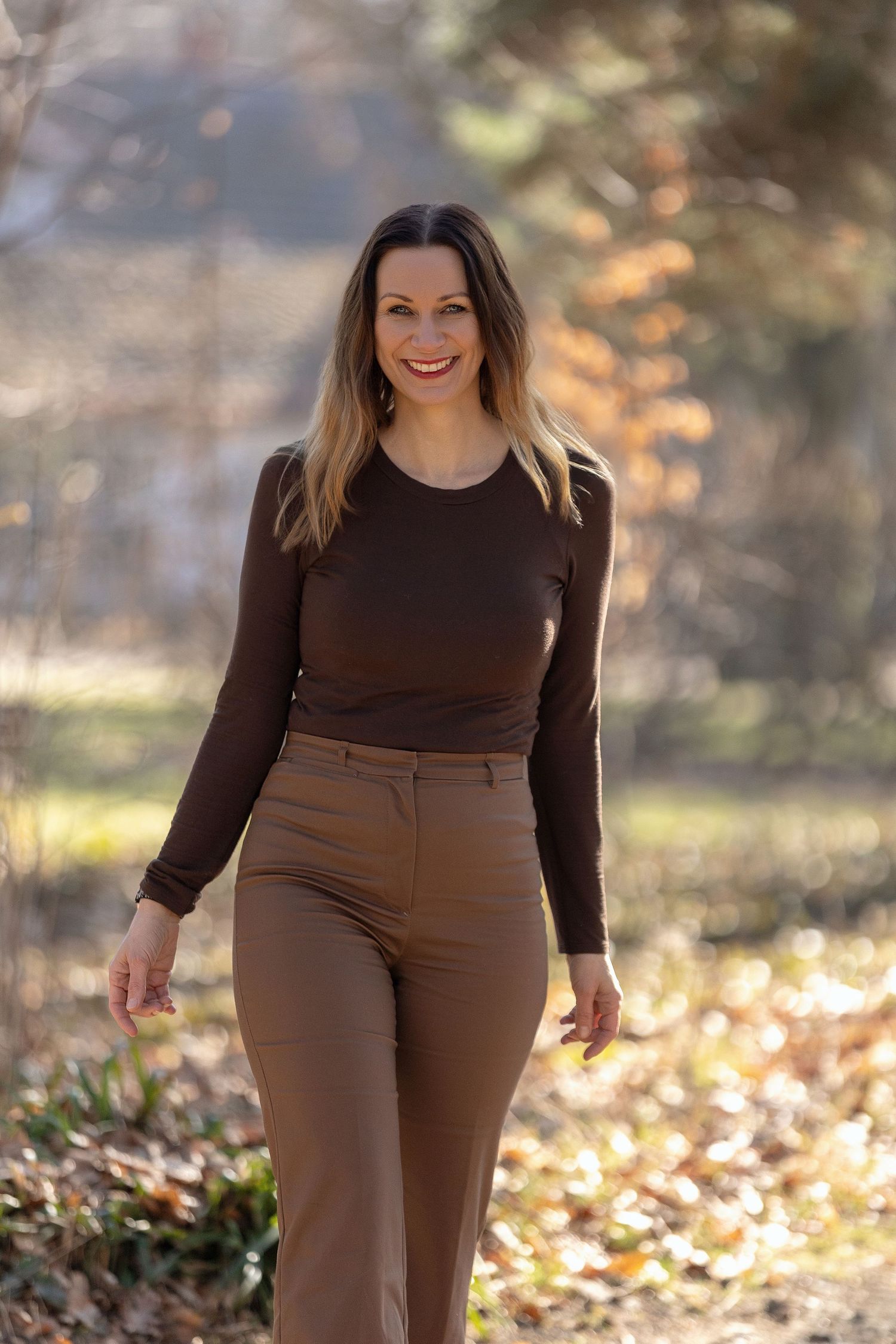Smiling woman in brown top and tan trousers walking outdoors in a sunlit autumn park.