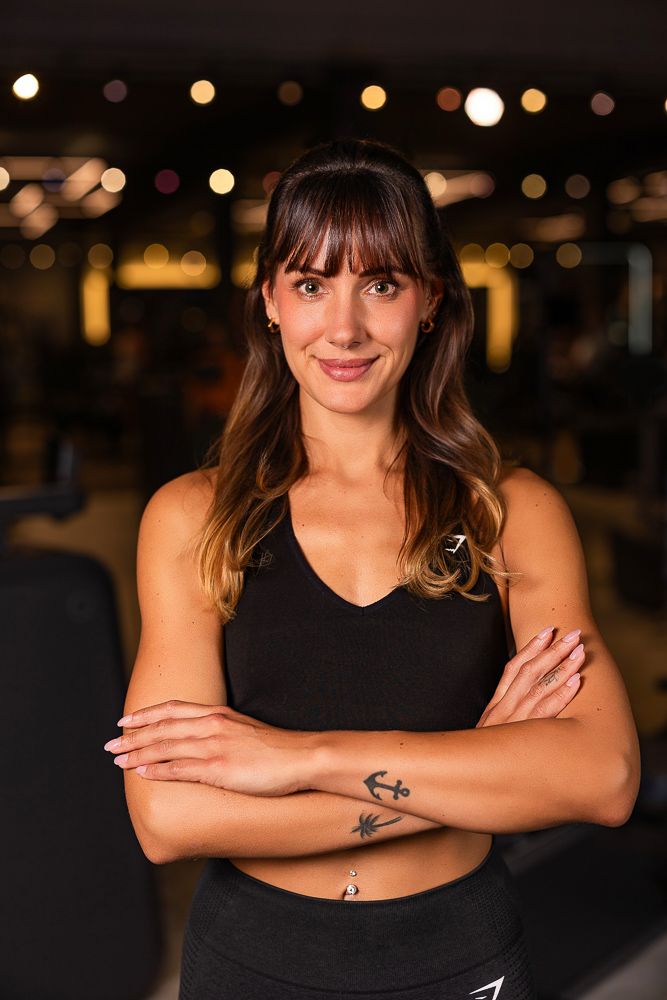 Fit woman with tattoos in black sports bra stands arms crossed smiling in gym.