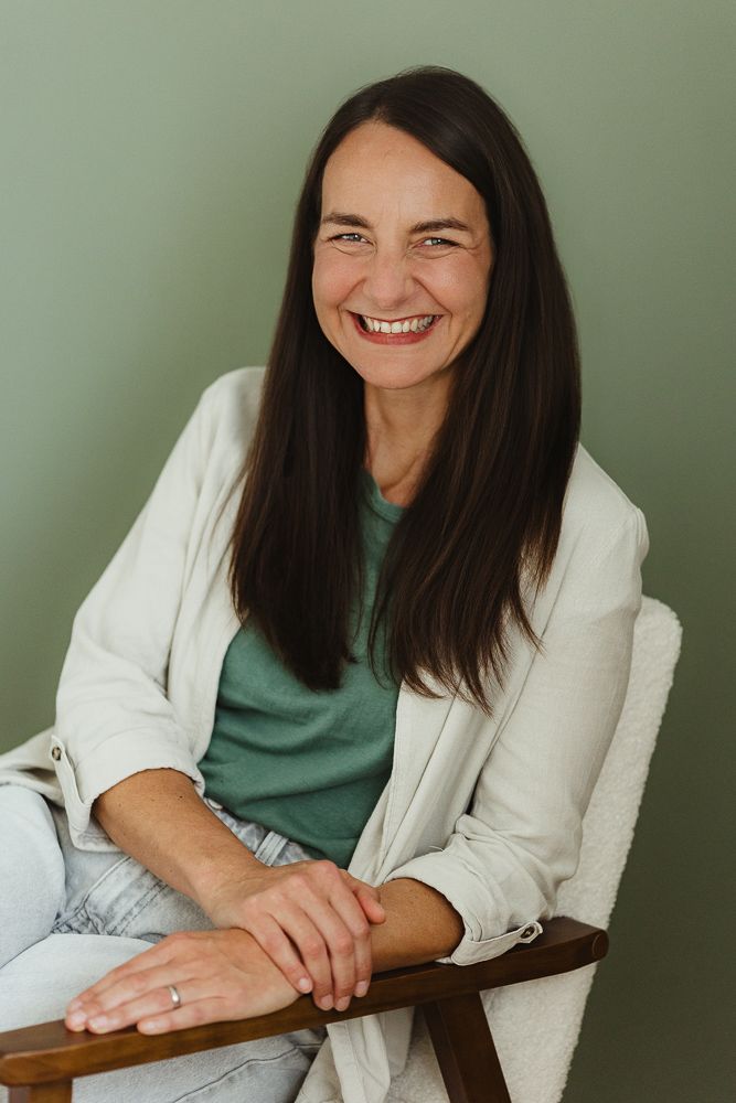Smiling woman with dark hair sitting in a wooden chair, wearing a green shirt and white blazer against a sage green background.