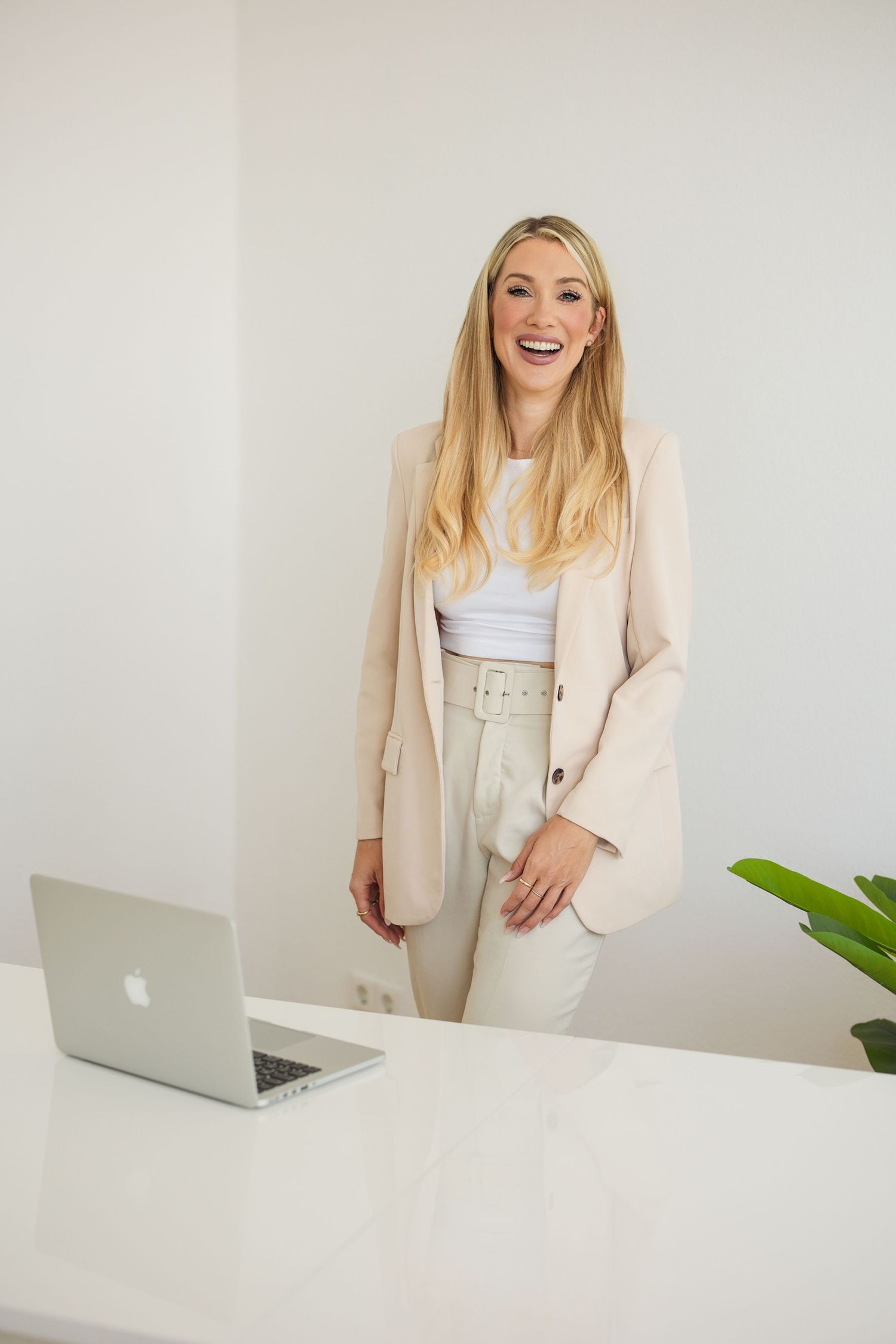 Professional woman in cream colored blazer and pants smiles while standing at white desk with laptop.