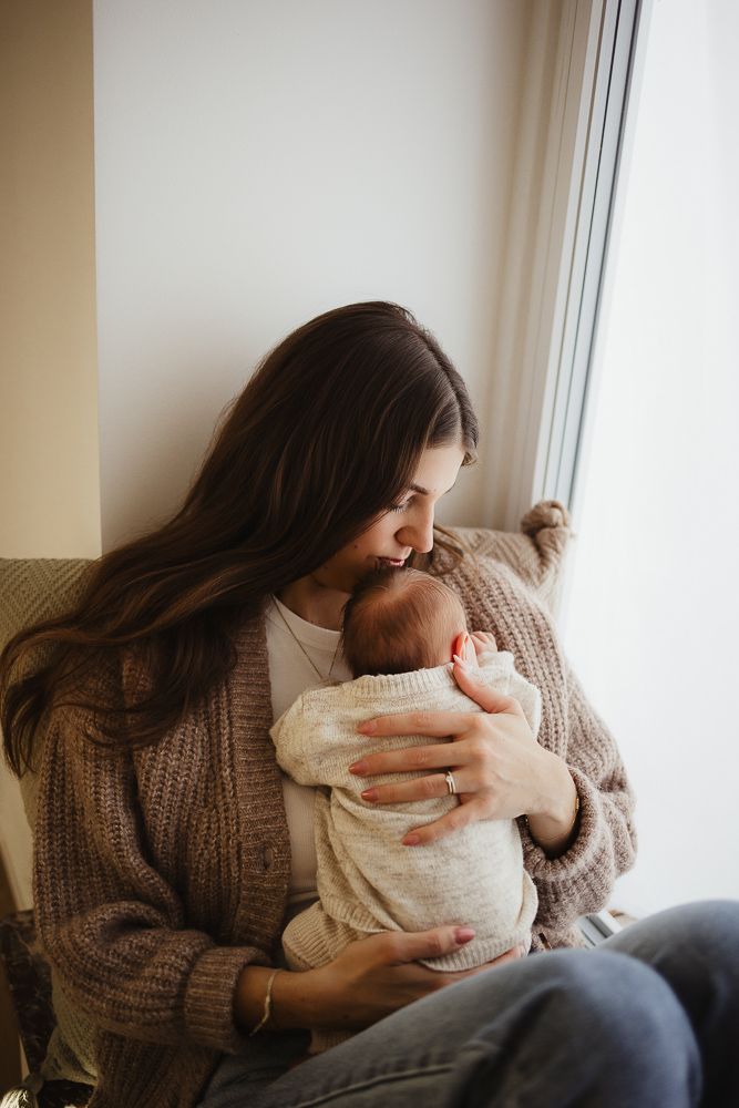 A young mother with long brown hair gently kisses her newborn baby while sitting by a window in a cozy knit cardigan.
