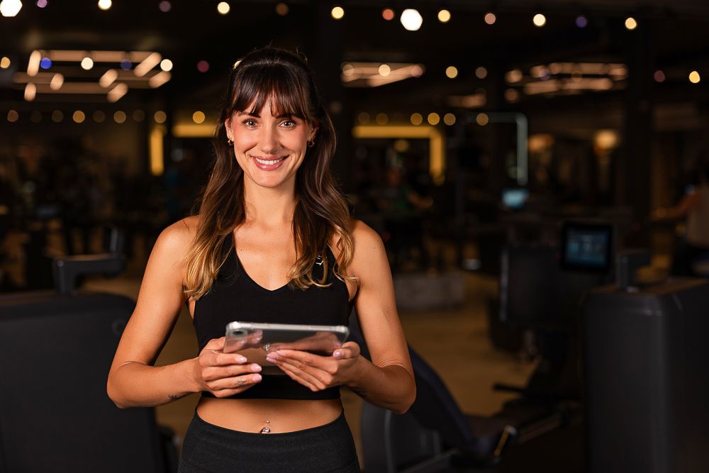 Smiling female fitness trainer holding a tablet in a dark gym environment wearing black athletic wear.