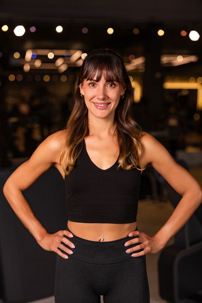 Confident woman in black sports bra and leggings posing with hands on hips in a gym setting.