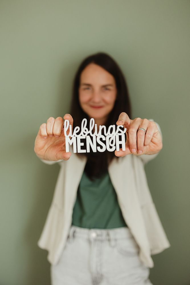 Woman holding a white decorative sign reading 'Lieblings MENSCH' against a sage green wall.