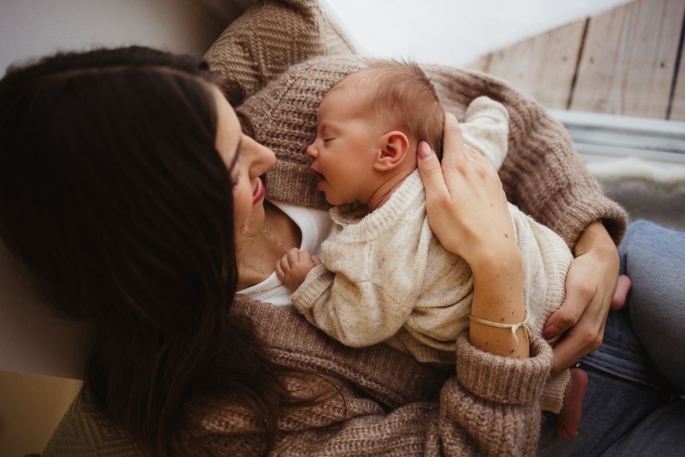 A mother in a cozy knit sweater tenderly holds her newborn baby, sharing a loving gaze in a warm, intimate moment.