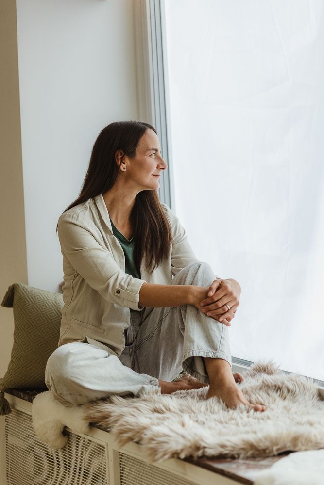 A contemplative woman sits on a window seat, hugging her knees, gazing thoughtfully outside in a cozy, light-filled room.