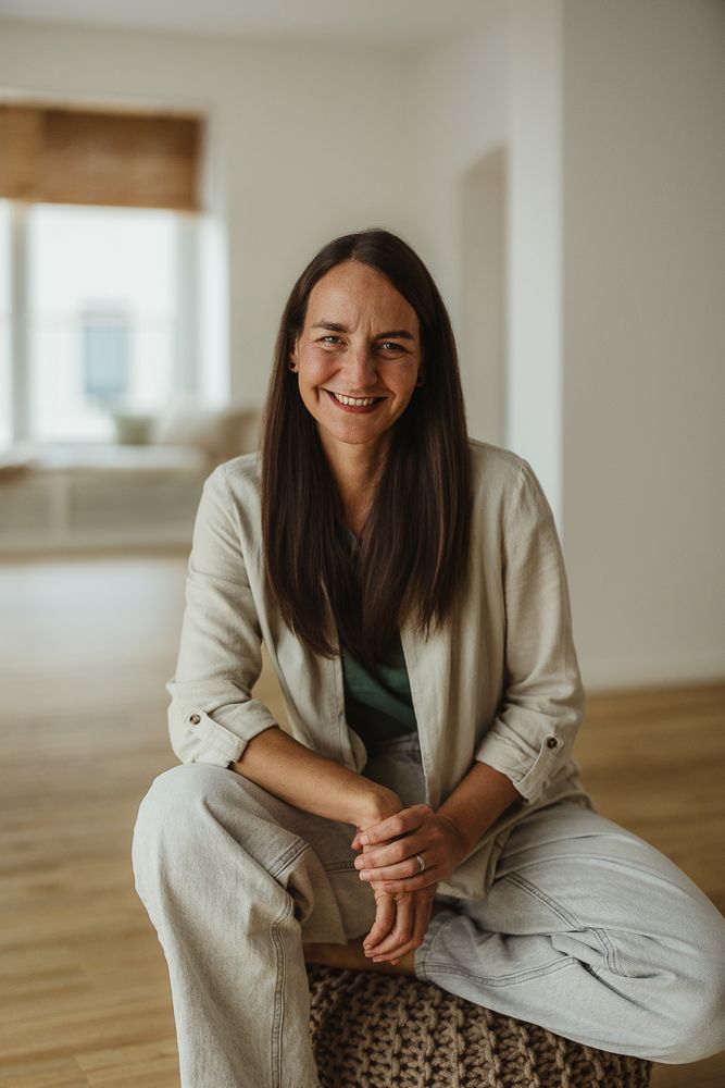 Smiling woman with dark hair wearing linen outfit seated in bright minimalist room with wooden floors.