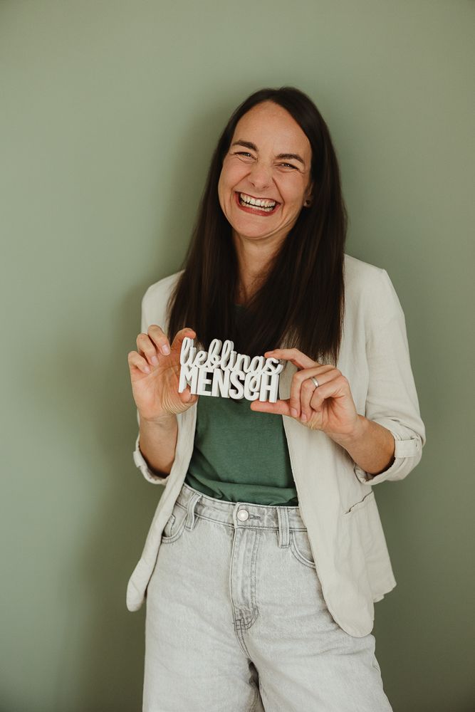 Laughing woman holding a white decorative sign reading Lieblingsmensch, wearing a blazer and jeans against a green wall.