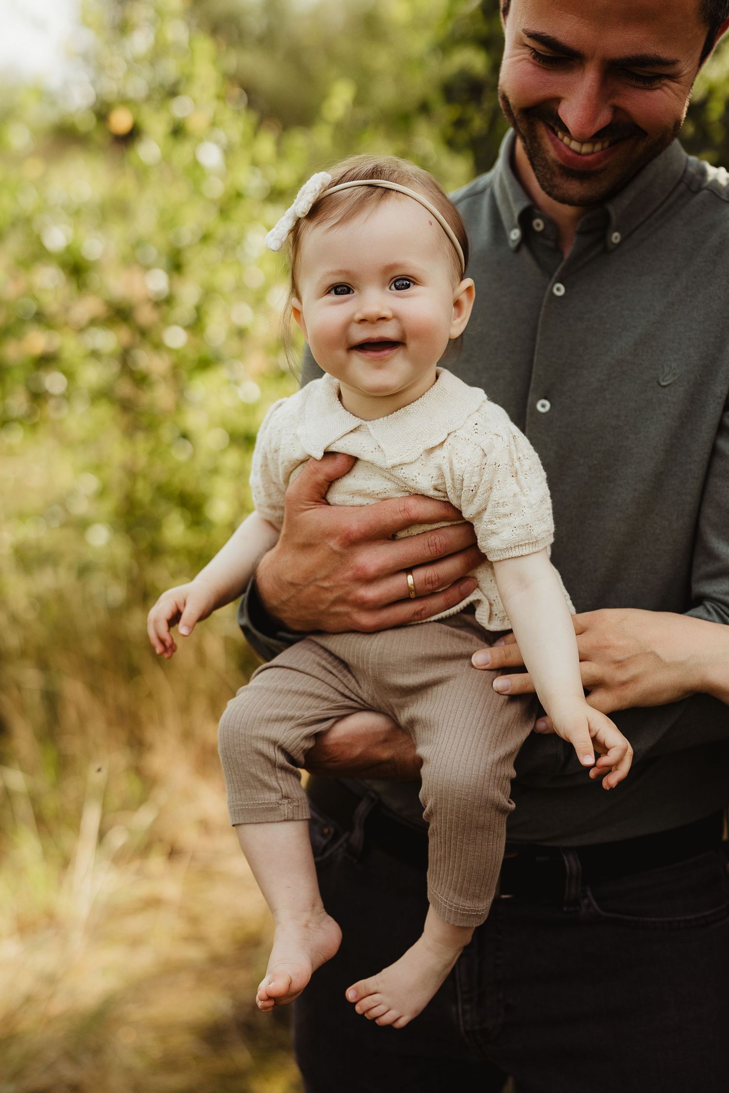 Happy baby in light colored outfit being held up outdoors in natural setting.