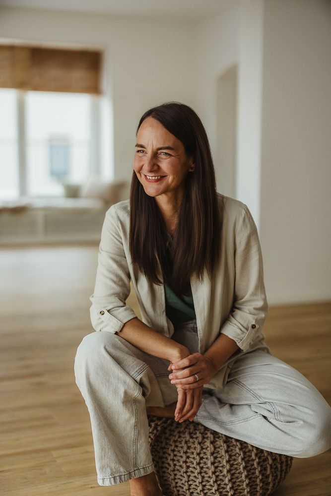 Smiling brunette woman sitting on a woven pouf in a bright, minimalist room wearing a linen jacket and jeans.