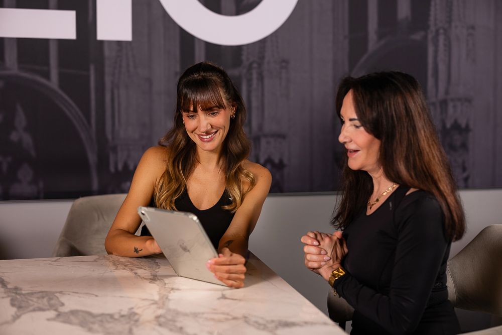 Two women smiling and reviewing content on a tablet at a marble table in a modern office setting.