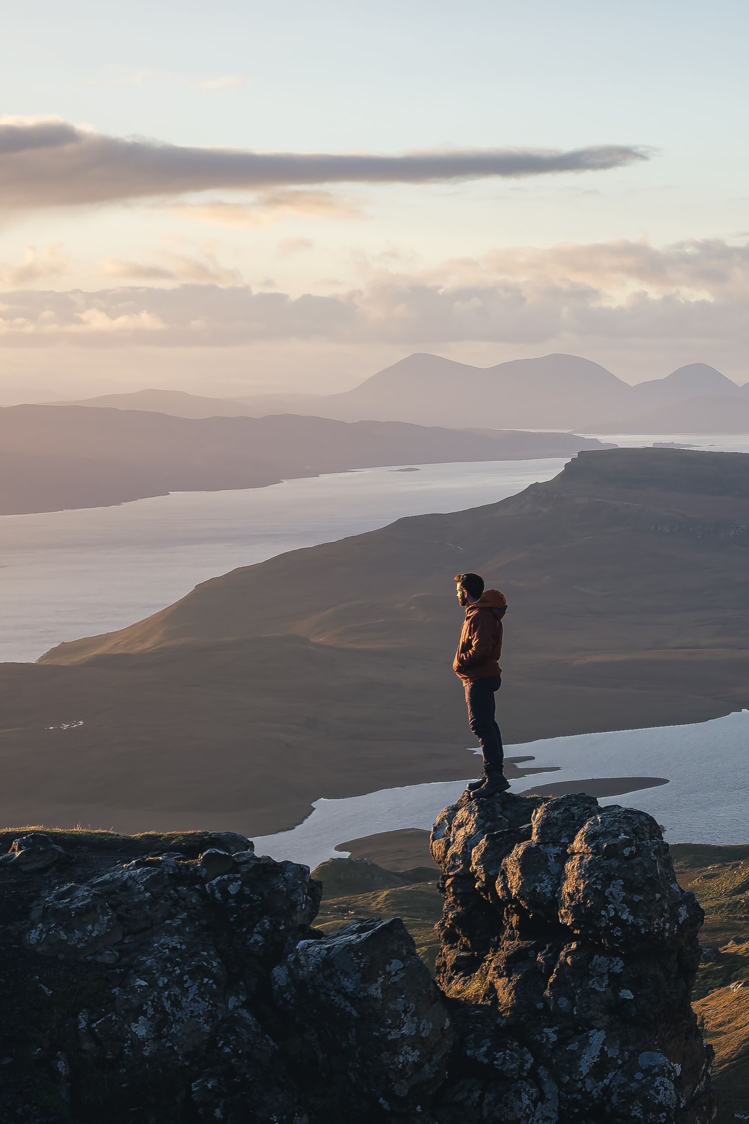 Randonneur en doudoune technique orange sur un pic rocheux, surplombant les lochs de l'île de Skye au coucher du soleil.
