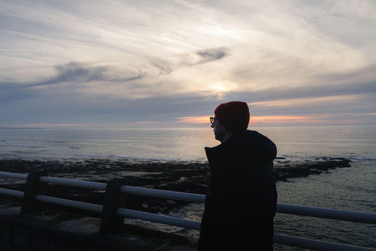Silhouette d'homme au bonnet rouge contemplant l'océan au coucher du soleil depuis une rambarde. Ambiance calme et art de vivre côtier.